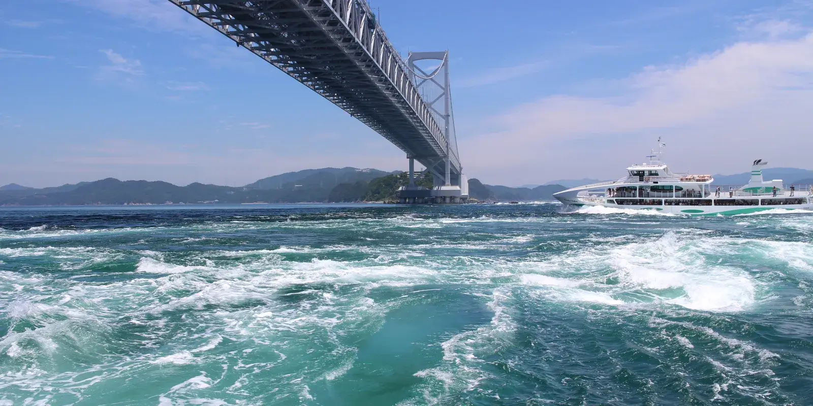 A tour boat passing beneath the Onaruto Bridge alongside churning turquoise whirlpool currents in the Naruto Strait