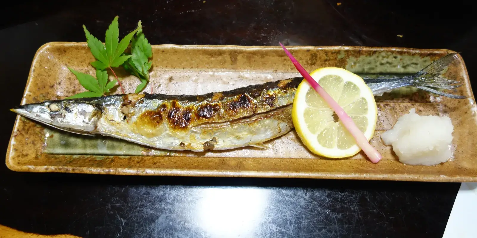 A whole salt-grilled sanma on a long ceramic plate with a lemon slice, grated daikon and a green maple leaf garnish