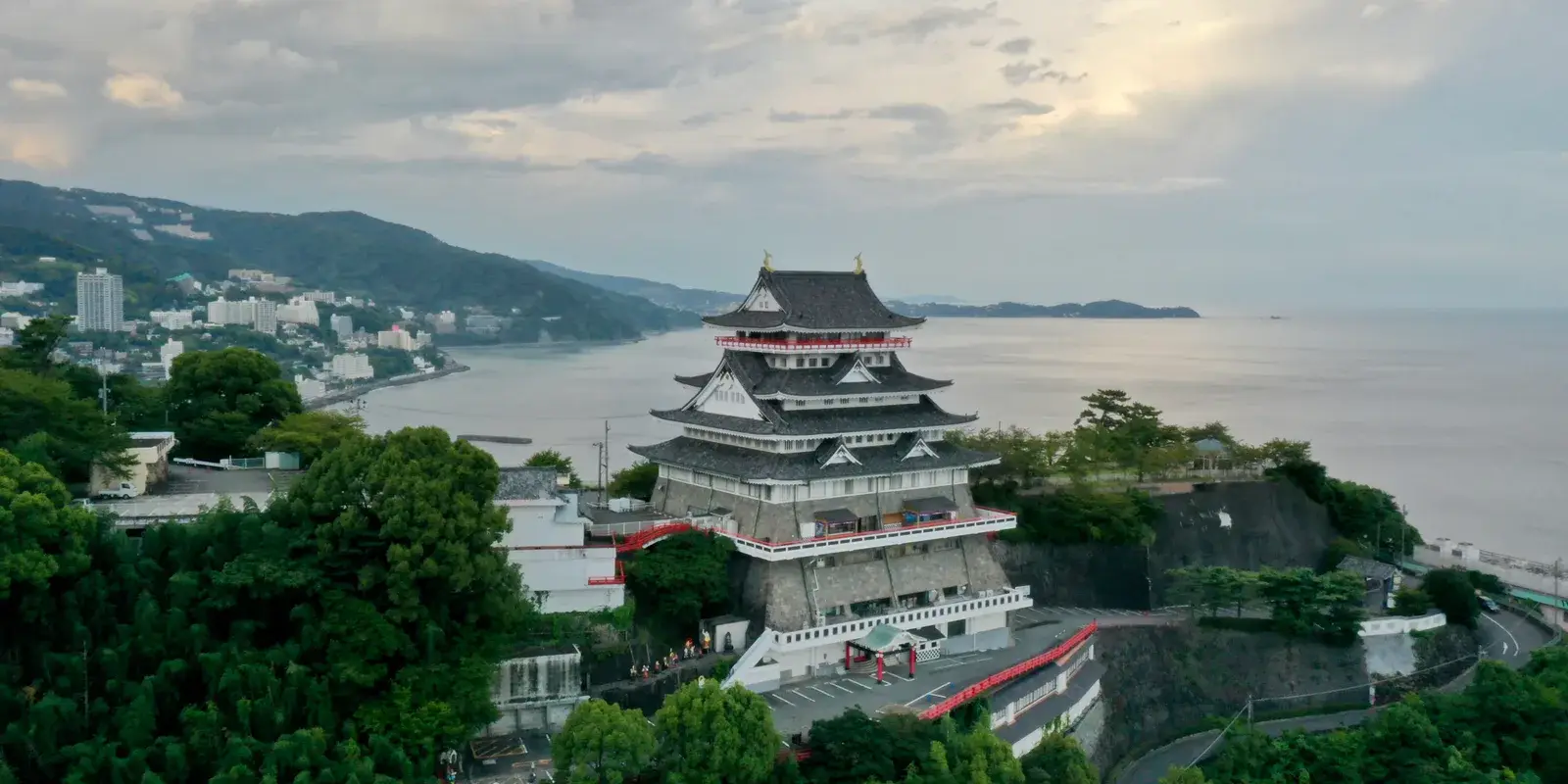 Aerial view of Atami Castle's tiered keep on a cliff above Sagami Bay, with the city and hills behind it