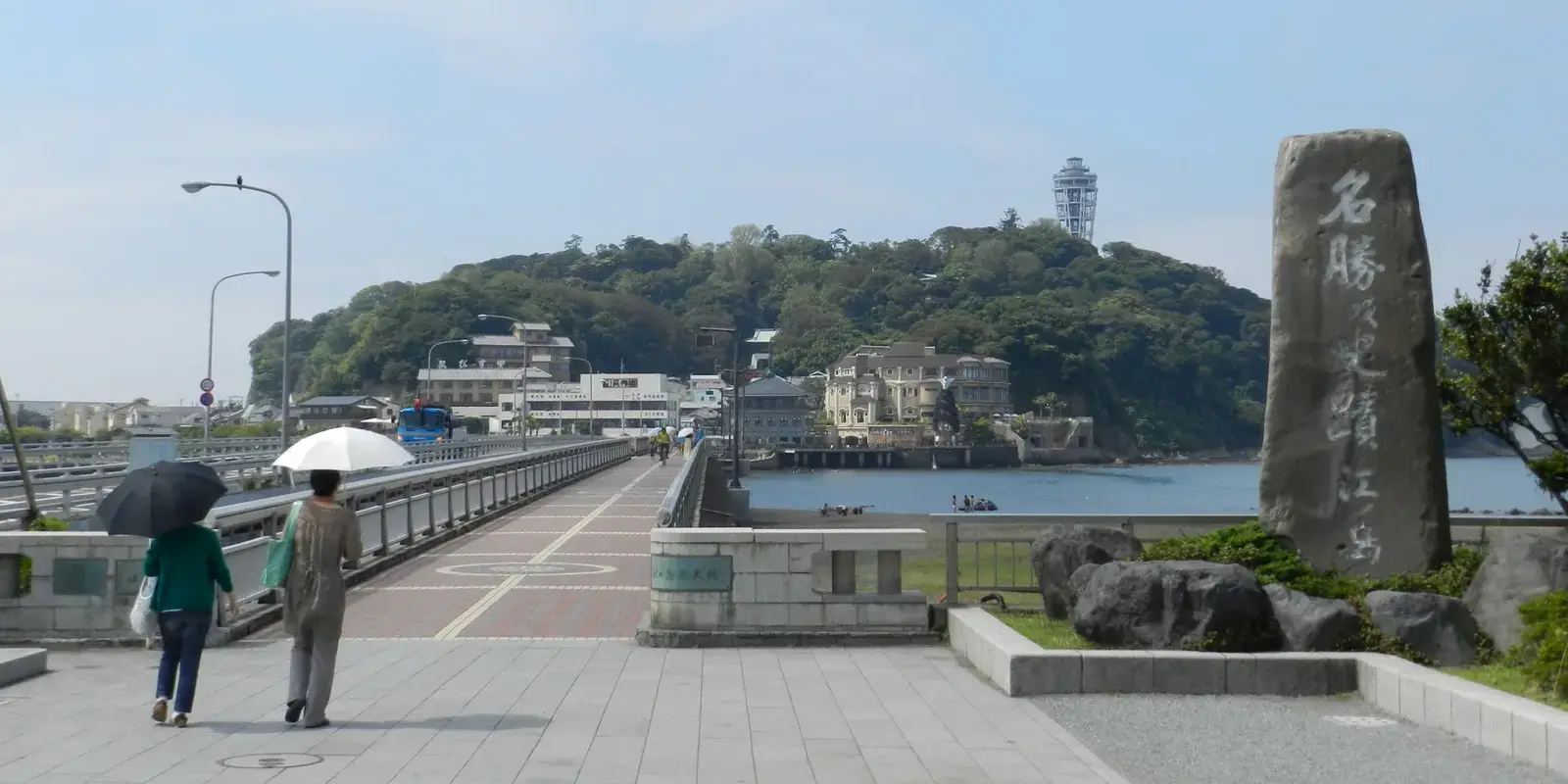 Two visitors with umbrellas crossing the bridge toward Enoshima island, with the Sea Candle tower visible on the wooded hill