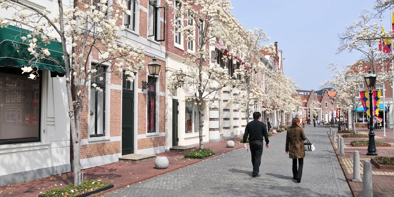 A couple walking down a magnolia-lined cobblestone street between Dutch-style brick buildings at Huis Ten Bosch