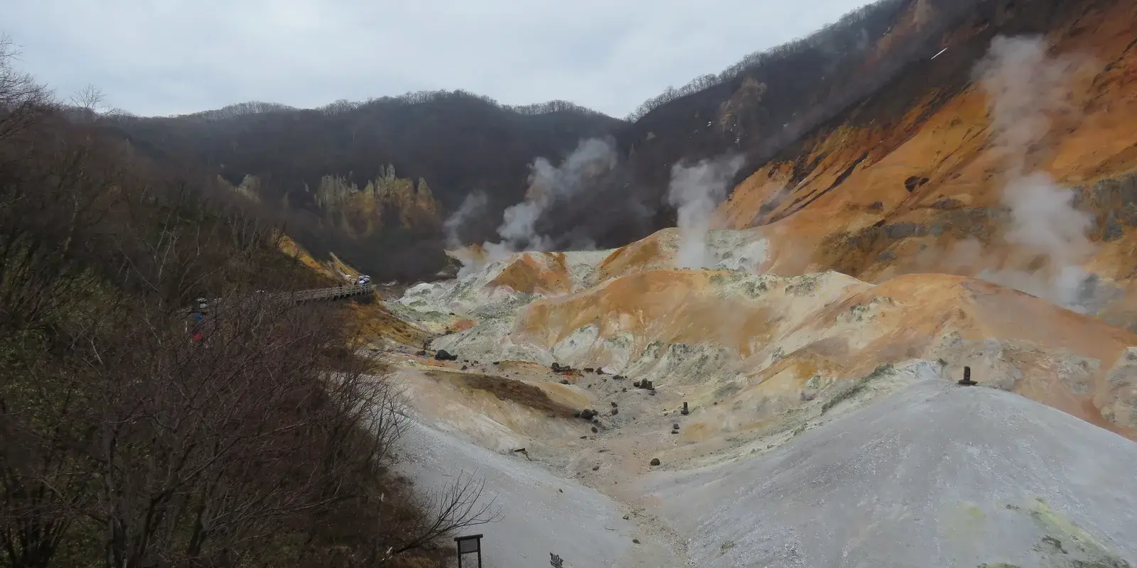 A wide view of Noboribetsu Jigokudani, its steaming valley of orange and grey volcanic rock ringed by forested hills