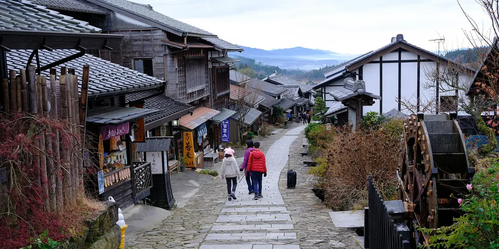 The stone-paved slope of Magome-juku descending between wooden inns toward the Kiso valley, with a water wheel turning at the roadside