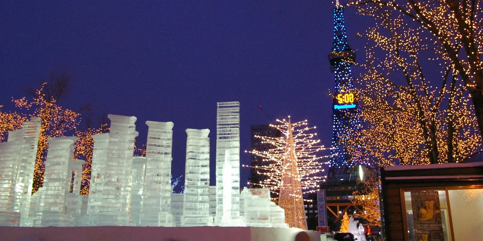 Ice sculptures glowing at dusk in Sapporo during the Snow Festival, with the illuminated Sapporo TV Tower and lit trees in the background