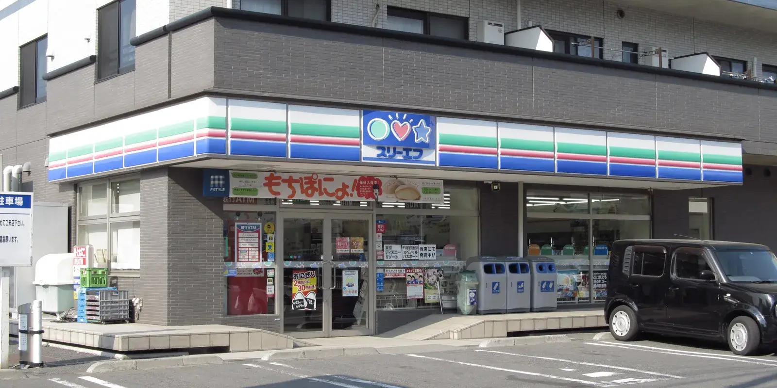 A Japanese convenience store (konbini) storefront in daylight with recycling and trash bins lined up by the entrance