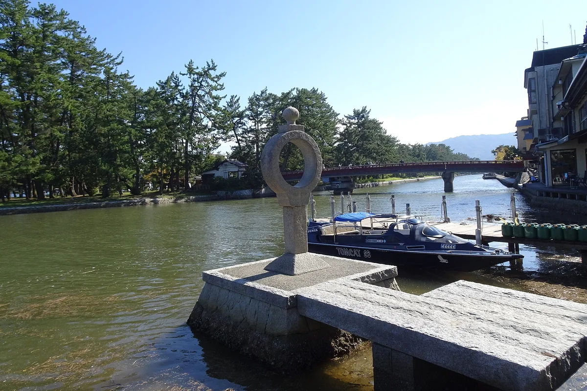 The red rotating bridge and stone Chie-no-wa ring monument at the Monju-side entrance to the Amanohashidate sandbar, with a tour boat docked alongside