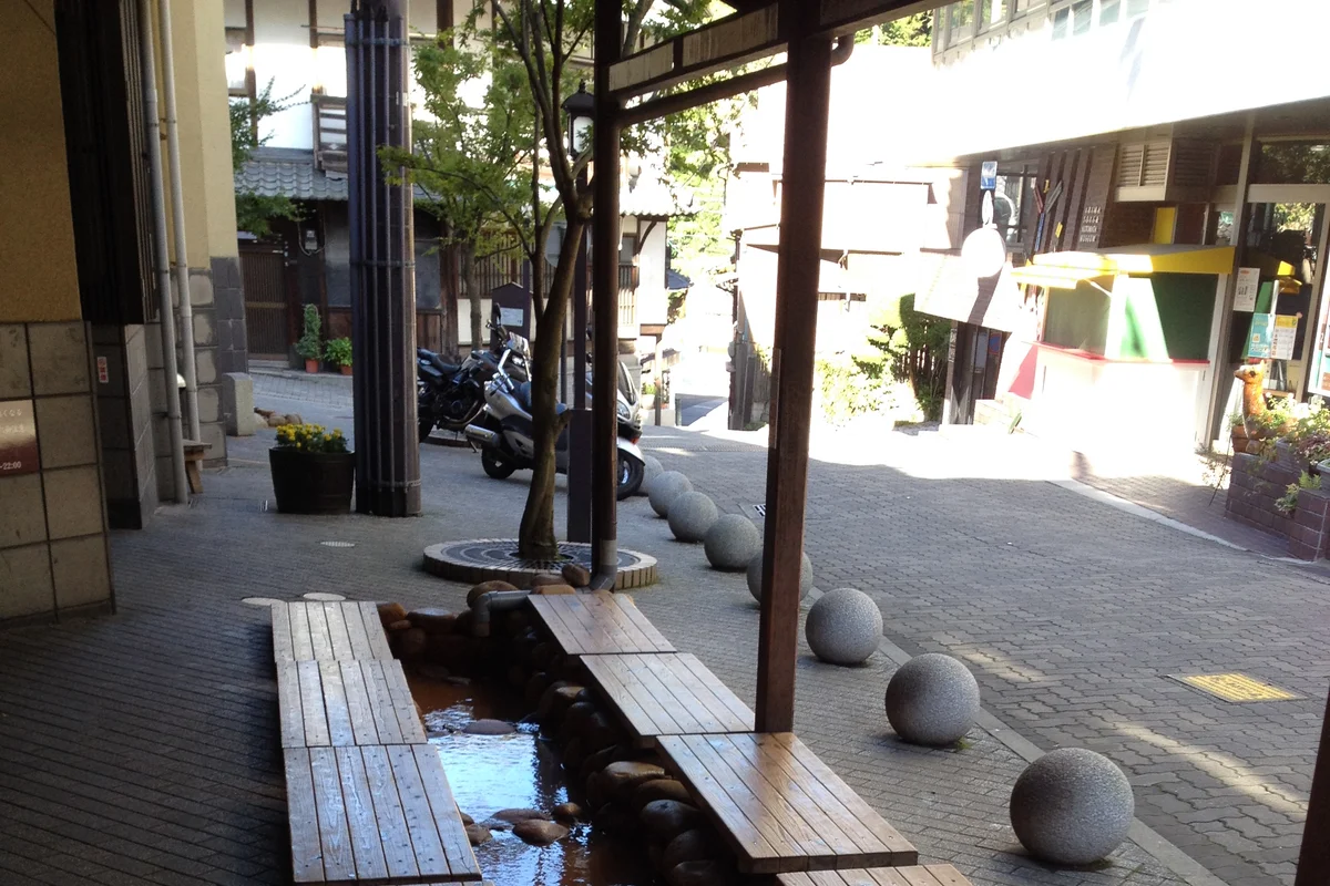 Wooden benches set over a shallow channel of reddish spring water at the free foot bath in Arima Onsen, beside a sloping street