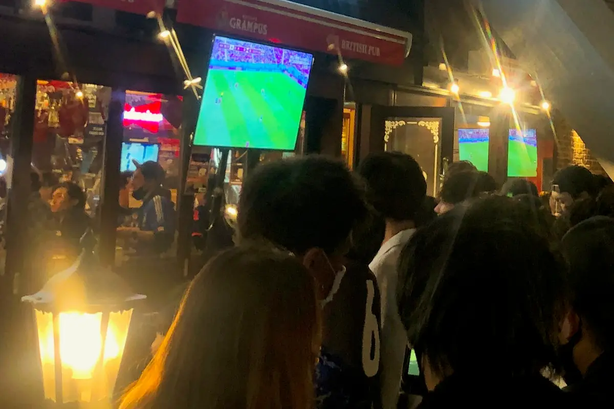 A night-time crowd seen from behind, standing outside a British-style pub in Japan and watching a football match on a large outdoor screen