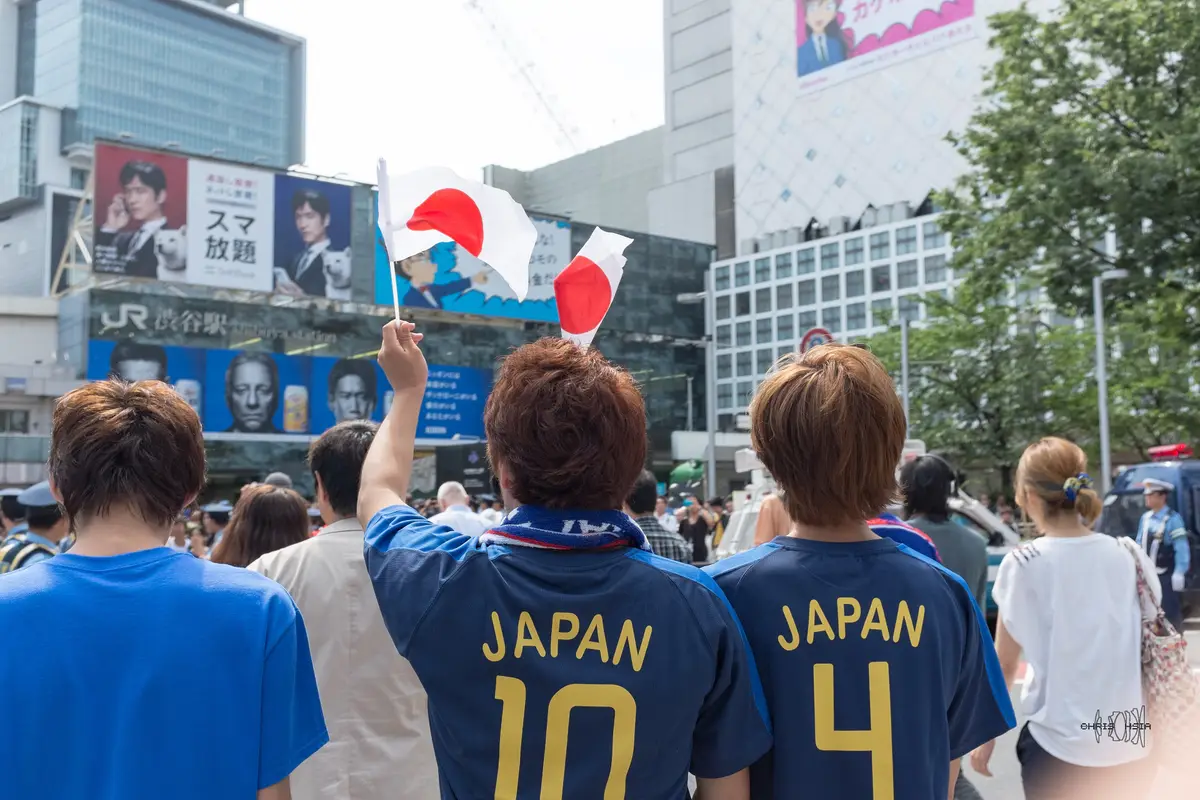 Supporters in blue Japan shirts seen from behind, holding small Japanese flags in front of the JR Shibuya Station sign, with police at the edge of the crowd
