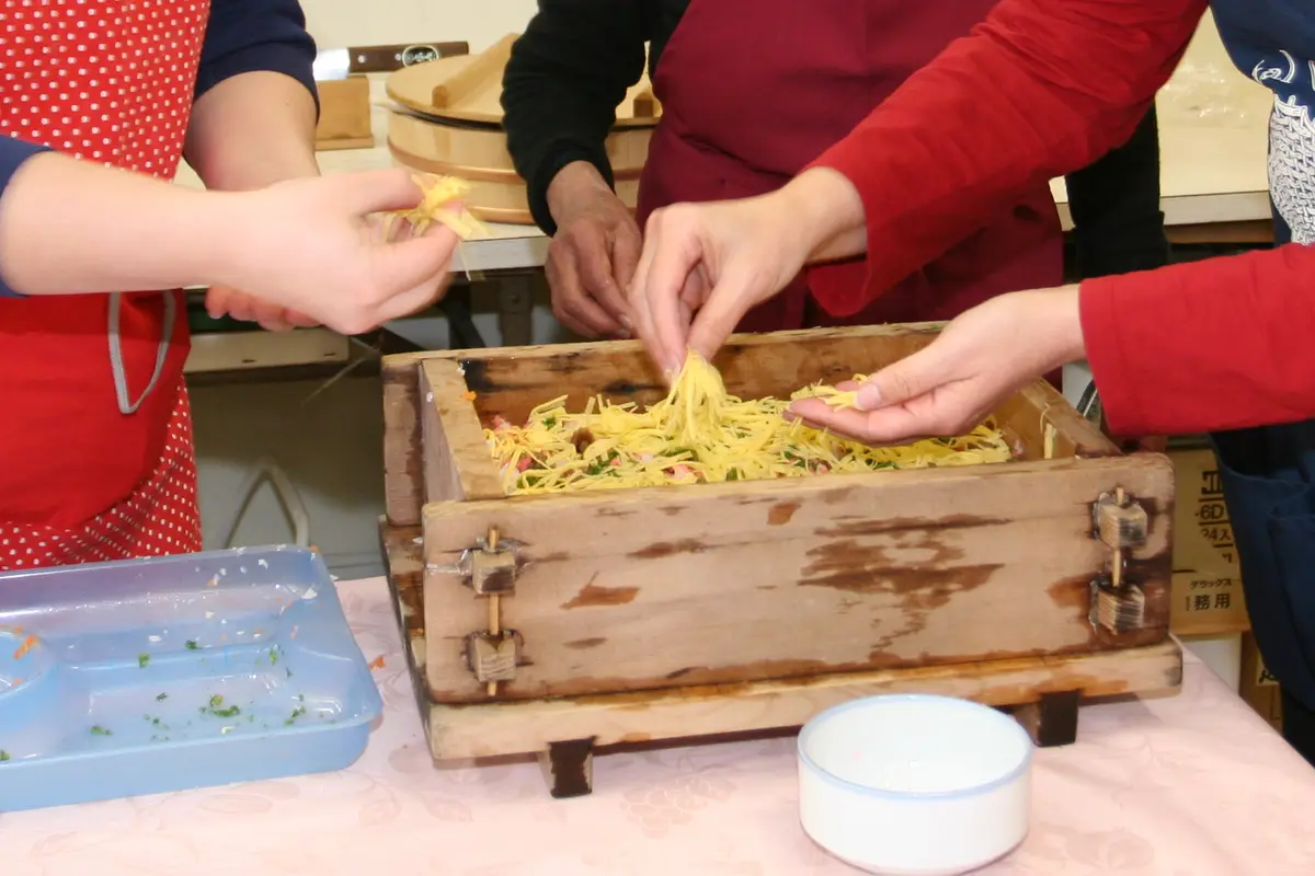 Hands scattering shredded egg over layered sushi rice inside a wooden pressing box at a cooking class in Japan