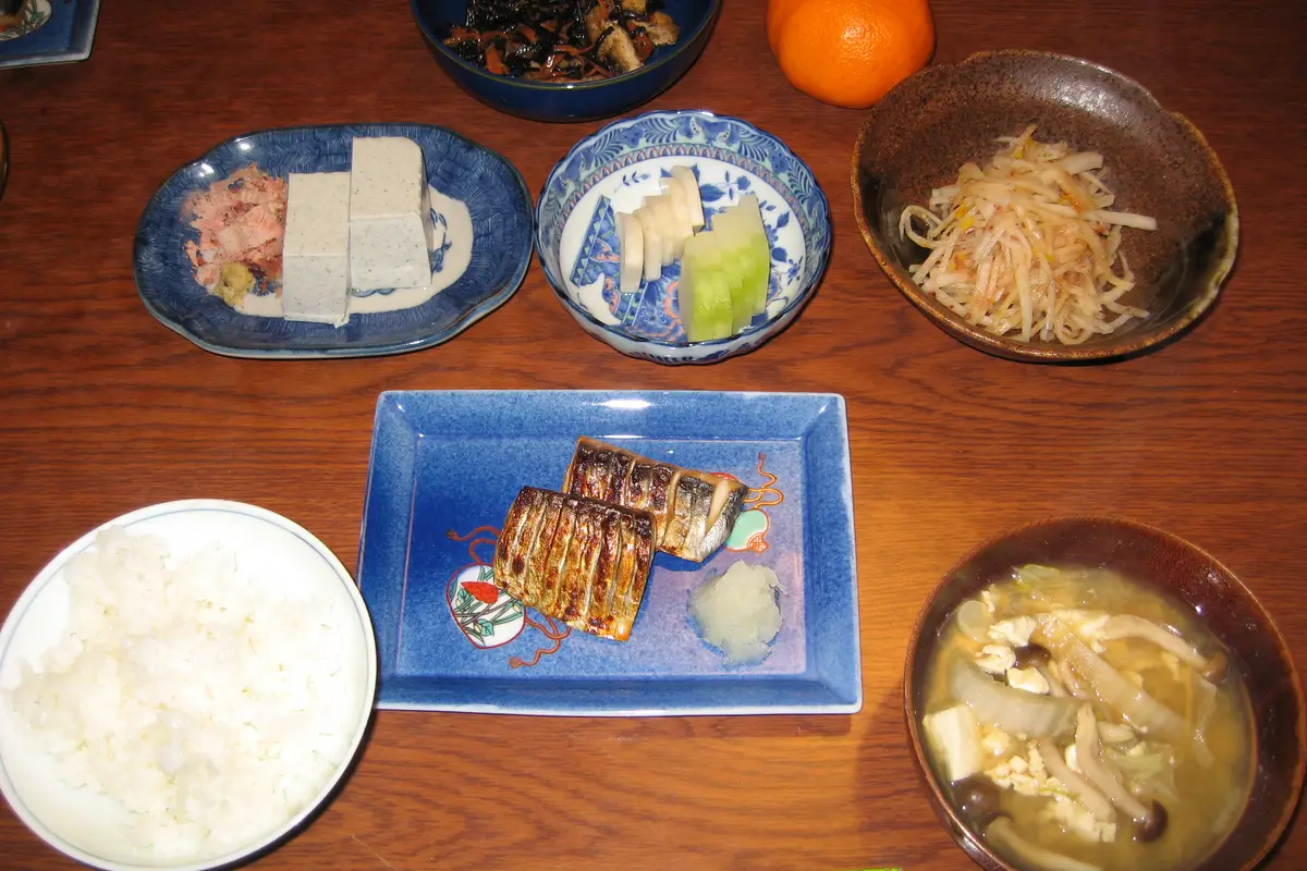 An everyday dinner in a Japanese home seen from above: a bowl of rice, miso soup, grilled fish, chilled tofu, pickles and a bean sprout side dish on a wooden table