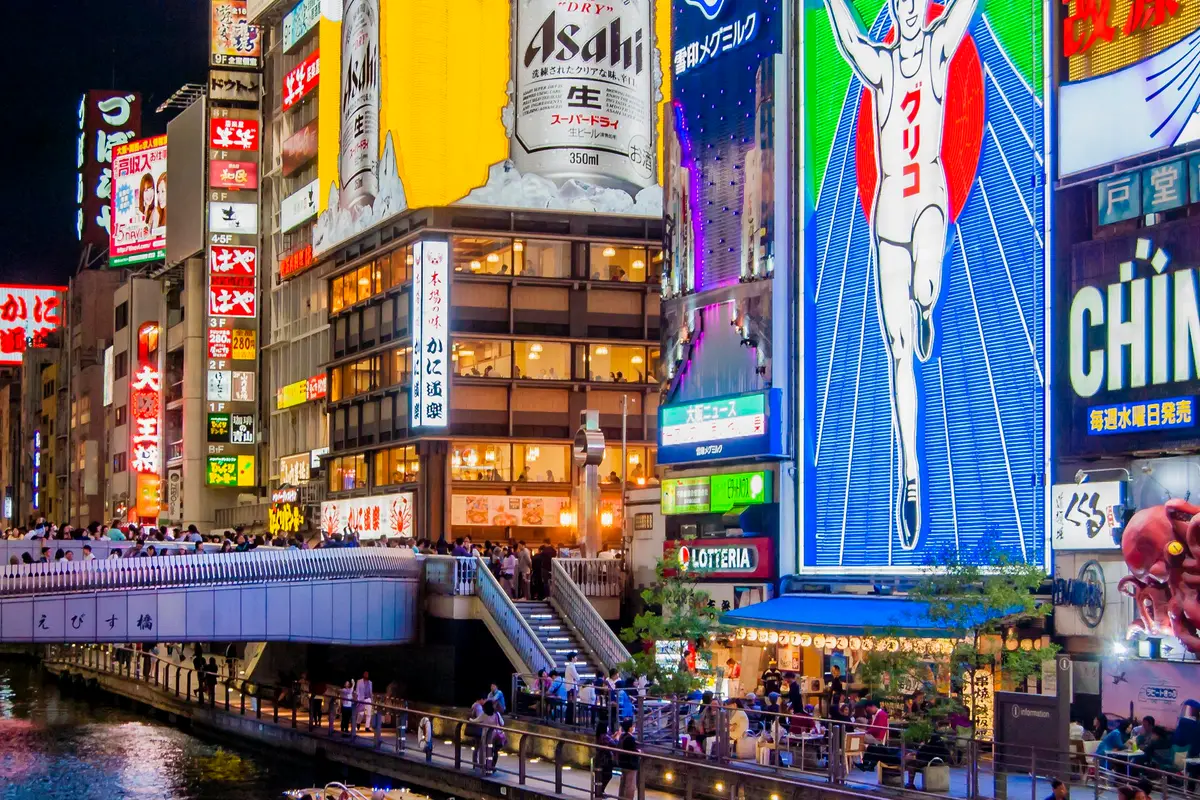 Ebisubashi bridge packed with people at night, beneath a wall of giant illuminated signs above the Dotonbori canal