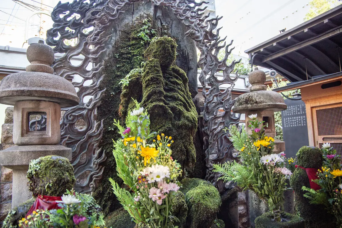 A moss-covered stone statue standing between two stone lanterns, with fresh flower offerings at its base, in Hozenji Yokocho