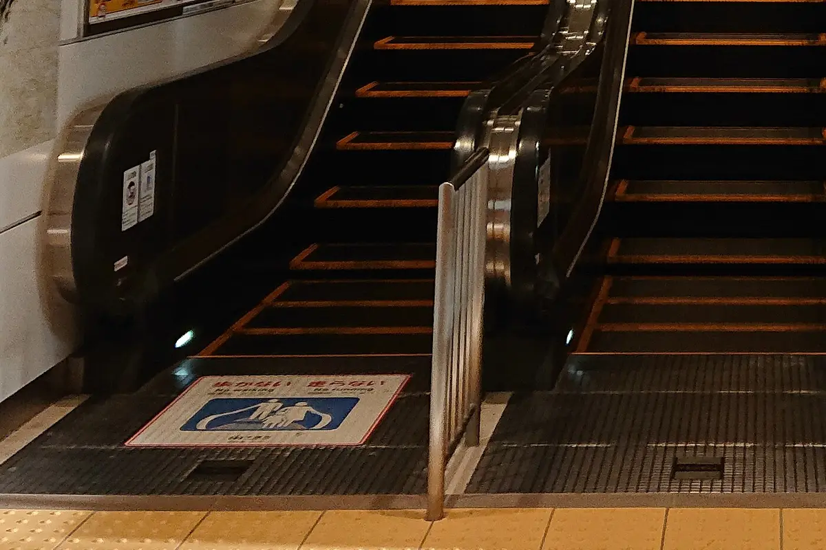 A floor sticker at the base of a station escalator showing two figures standing on the steps, labelled No walking and No running