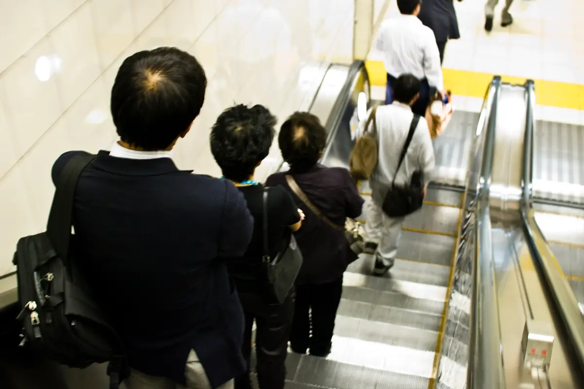 Passengers stand one behind another on one half of a station escalator while another person walks down the empty half