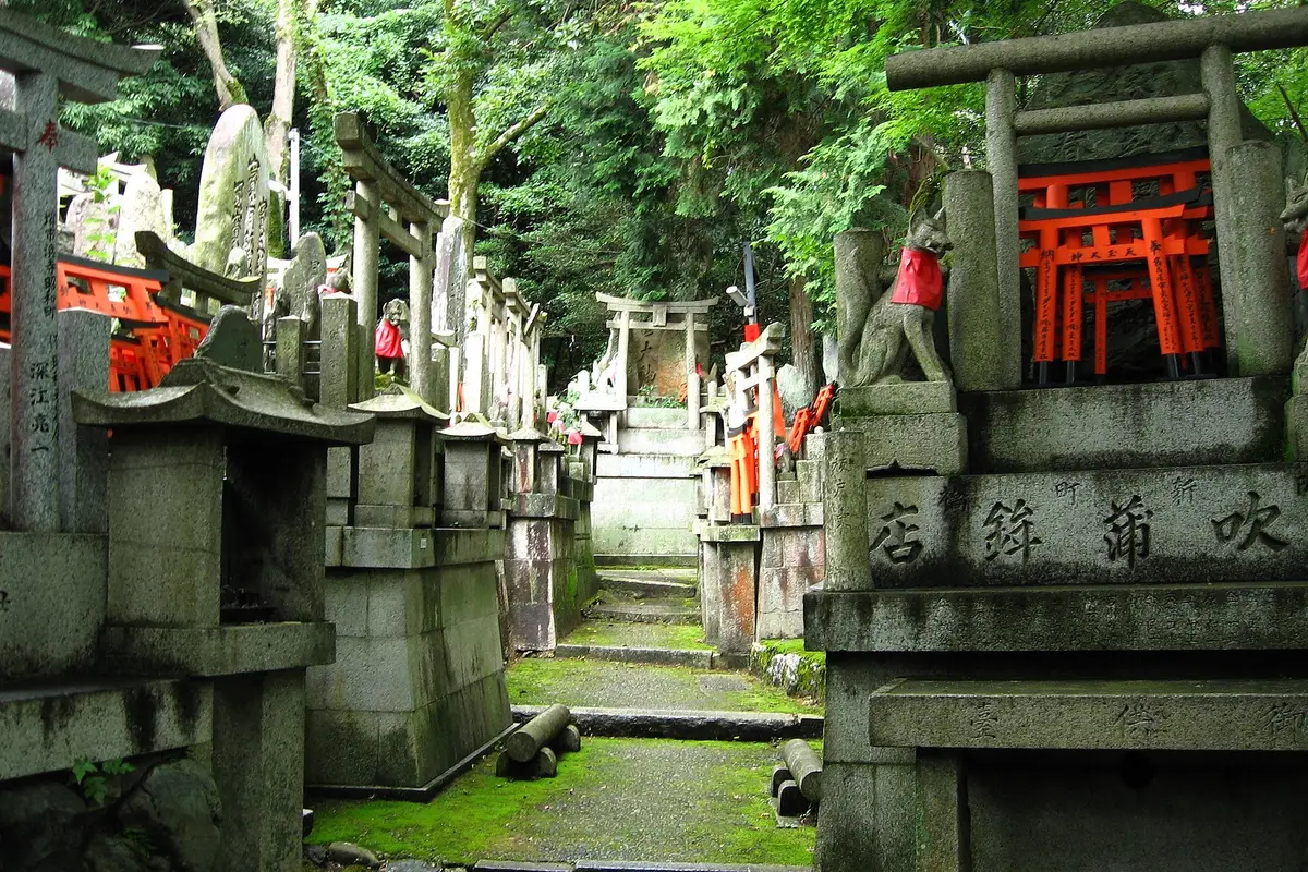 Small stone altars topped with miniature torii and fox statues in red bibs, along a mossy path in the woods of Mt Inari
