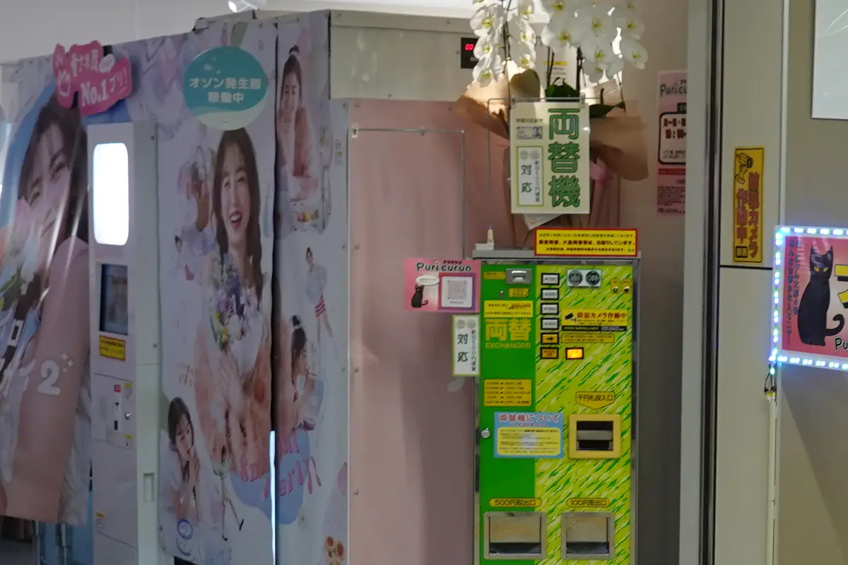 A purikura corner in Japan, booths wrapped in printed panels with a soft curtain and a change machine beside them