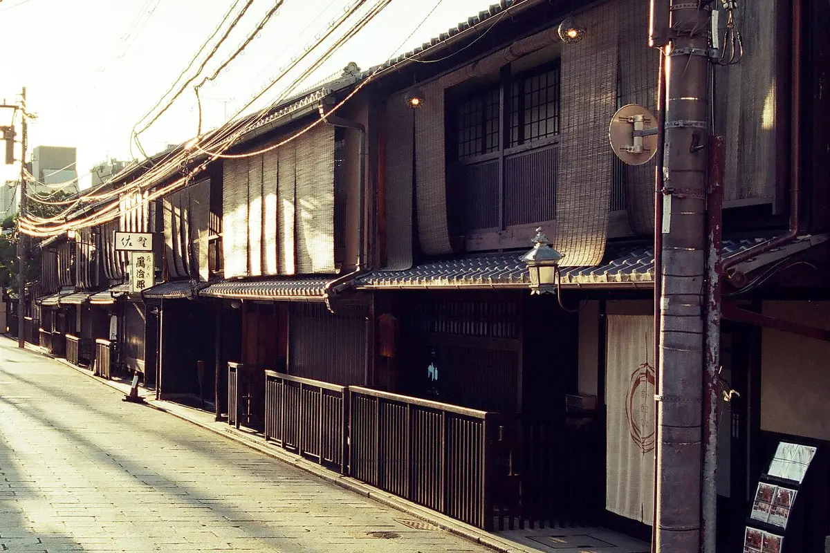 A row of two-storey wooden buildings in Gion, Kyoto, with reed screens, a hanging lantern and an empty stone-paved street in warm low light.