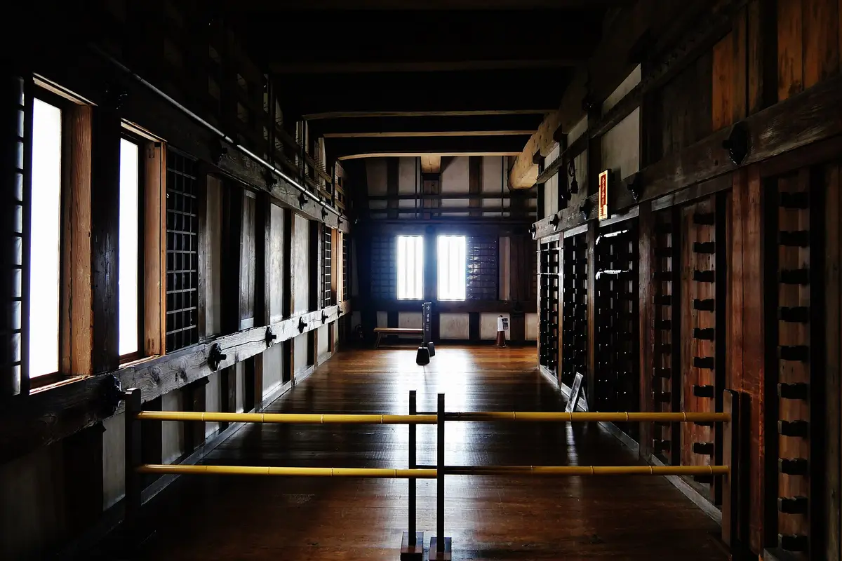 An empty upper floor inside the keep of Himeji Castle, with bare wooden floorboards, timber posts, and weapon racks along the wall