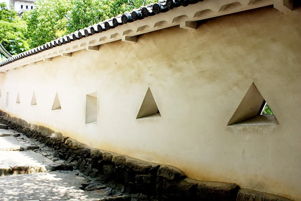 Triangular and square openings cut through the white plaster wall of Himeji Castle, above a stone footing beside the path