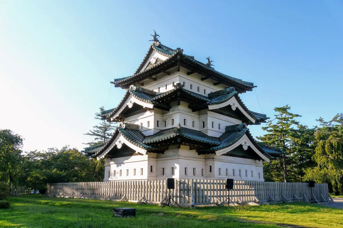 Seen across open grass, the small three-story wooden keep of Hirosaki Castle standing on a low temporary base behind a wooden fence