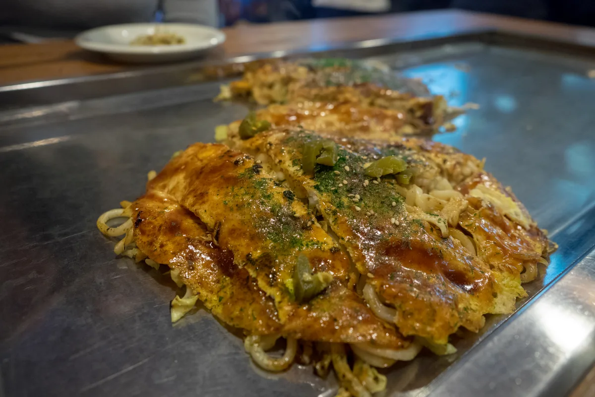 Hiroshima-style okonomiyaki being cooked on a flat iron grill, layered with noodles and cabbage