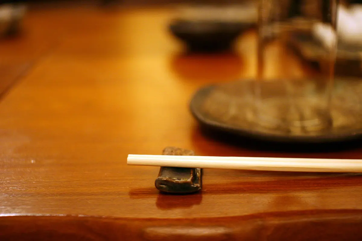A pair of chopsticks laid across a small ceramic chopstick rest on a wooden table, with a dish blurred behind
