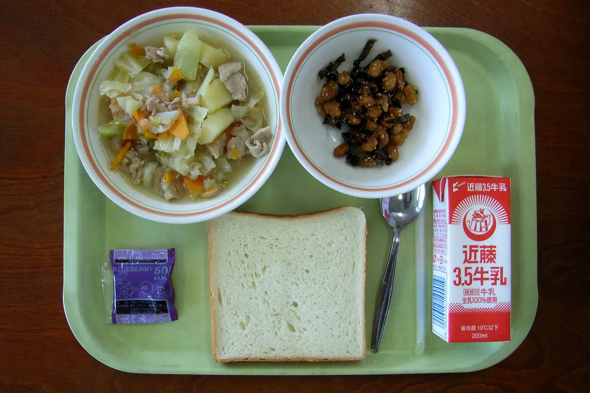 A Japanese elementary school lunch tray seen from above, with a bowl of meat and vegetable soup, a small dish of seasoned beans and seaweed, a slice of bread, a jam packet, a spoon and a carton of milk