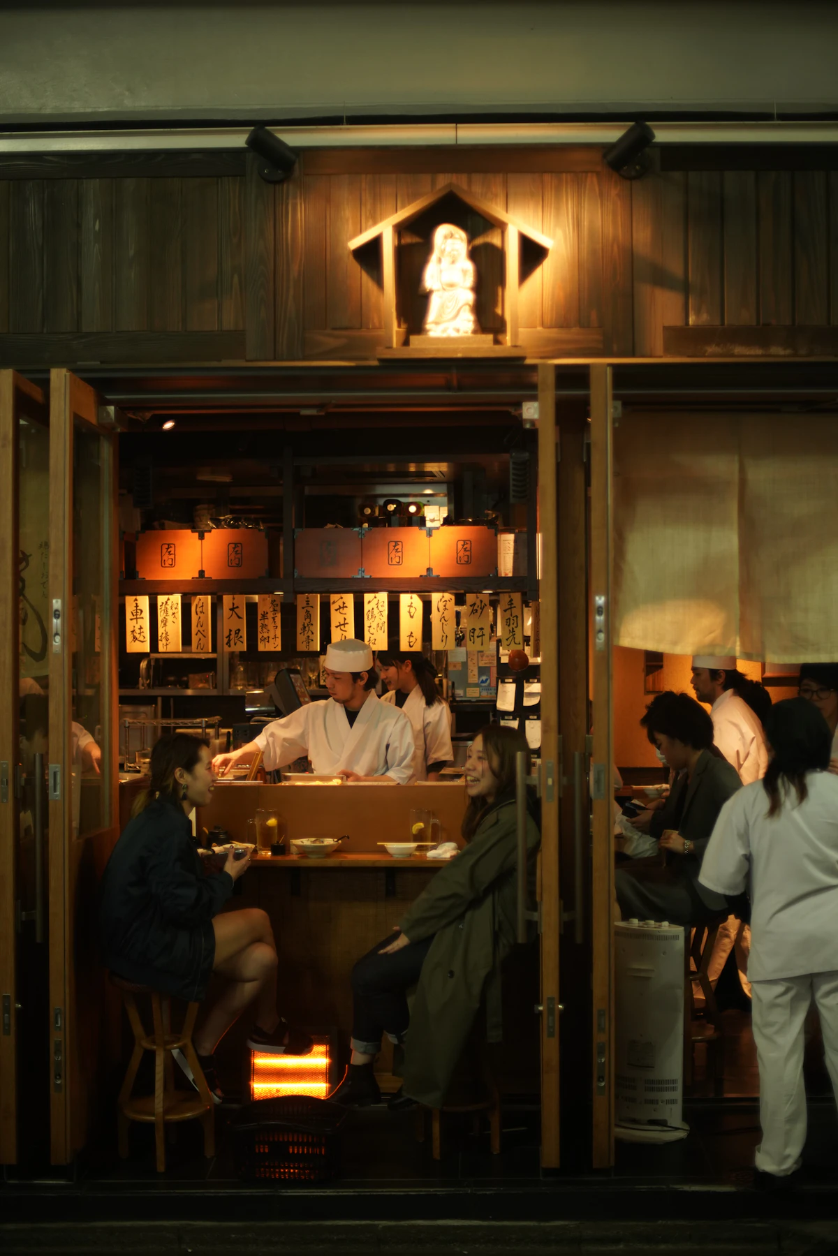 A chef and customers at an izakaya counter seen through the open doorway
