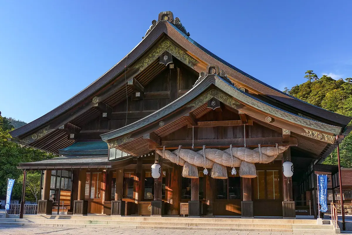 The worship hall of Izumo Taisha under a clear sky, its braided straw rope hanging beneath the deep wooden eaves