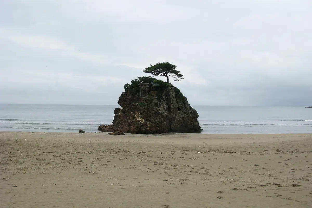 The wide dry sand of Inasa Beach, with a rock island carrying a small torii gate standing where the waves break