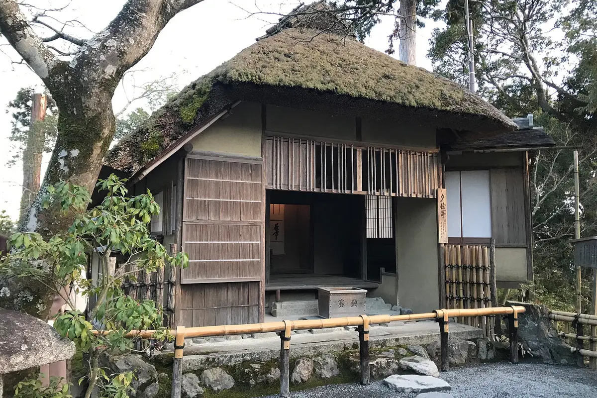 Sekkatei at Kinkaku-ji: a small tea house with a thick thatched roof and weathered wooden shutters, standing on a stone base beside the gravel path