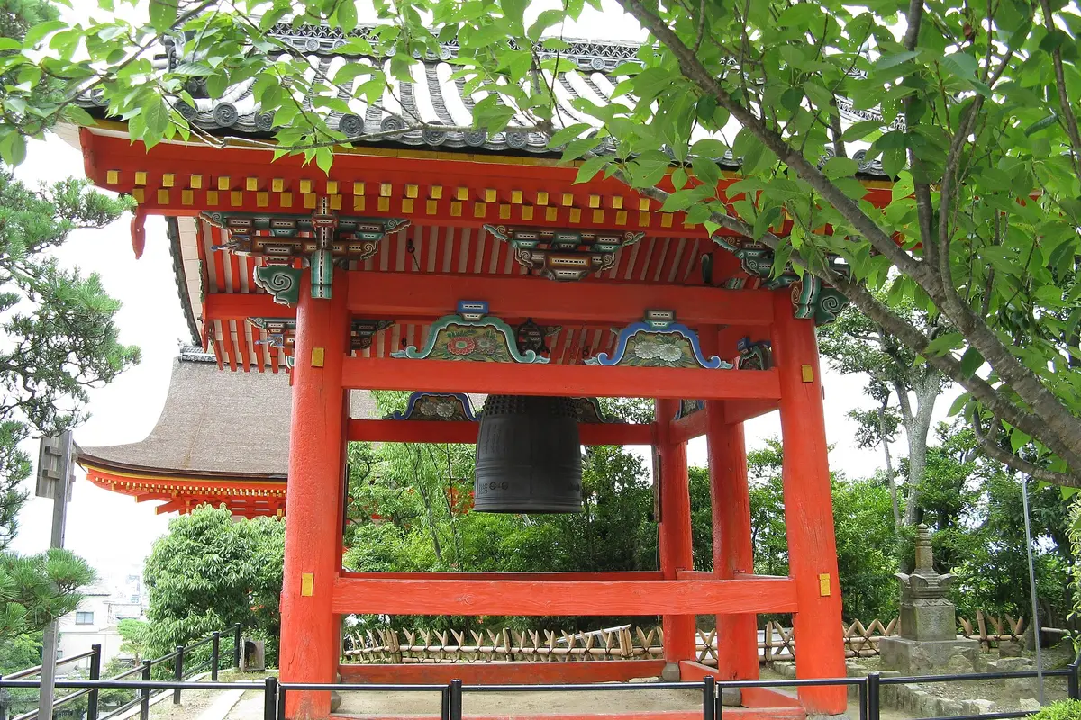 The vermilion bell tower of Kiyomizu-dera under green leaves, its great bronze bell hanging beneath painted brackets