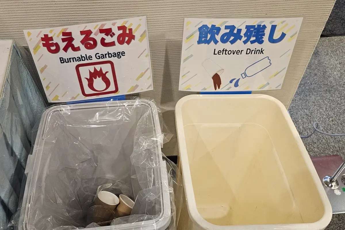 Two bins side by side with signs in Japanese and English reading burnable garbage and leftover drink, above a drawing of a paper cup and a plastic bottle being poured out