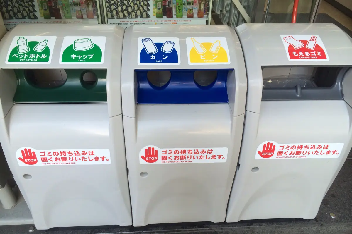 Close view of three sorting bins outside a Japanese store, with openings labeled for PET bottles, caps, cans, glass bottles and burnables