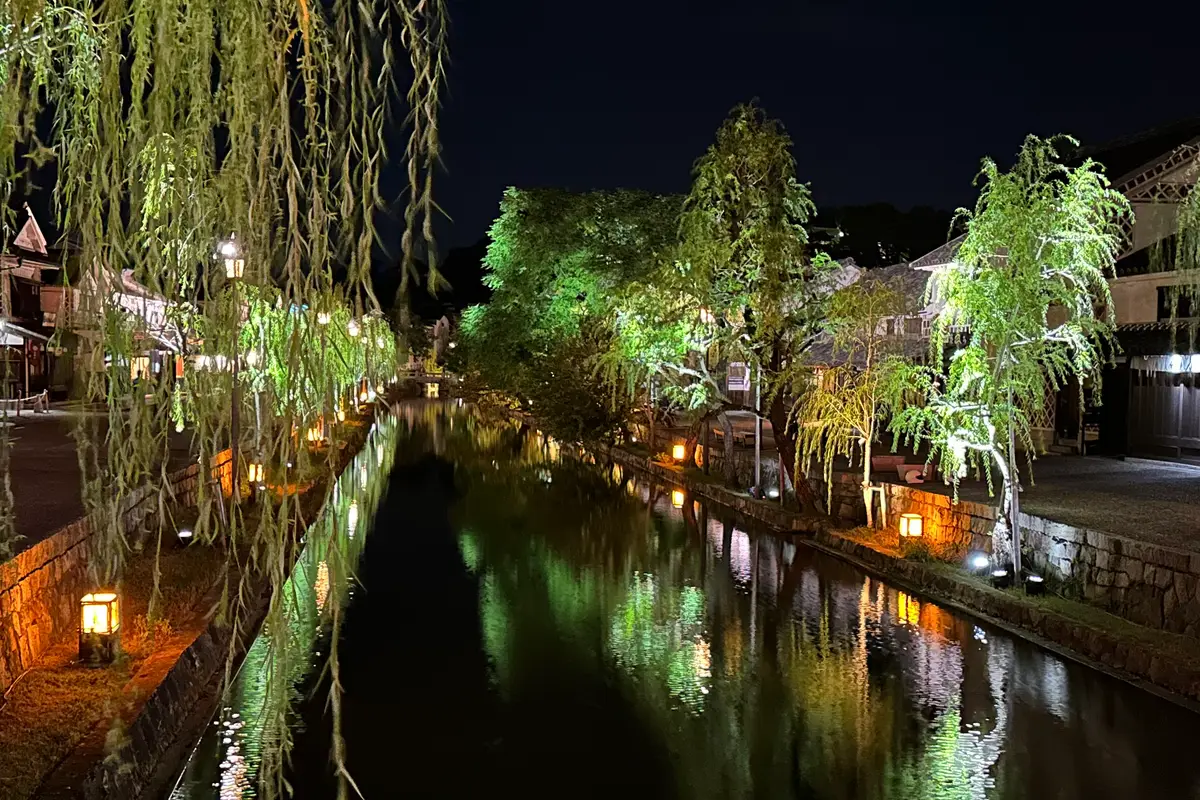 Seen from a bridge after dark: floodlit willows and lantern light reflected in the still water of the Kurashiki River, with an empty walkway along the stone banks