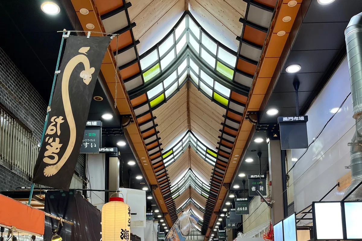 Looking up at the long wooden roof and skylights of Kuromon Market's covered arcade, with a shop banner and a paper lantern hanging below