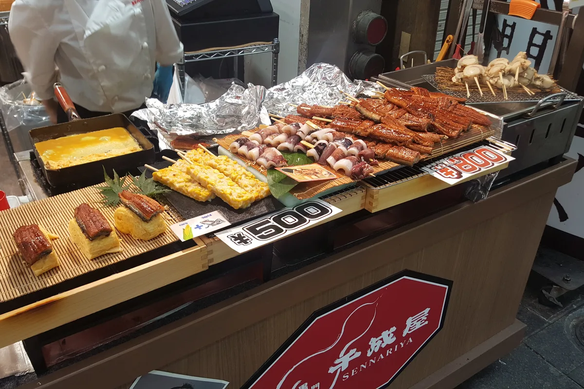 A seafood stall at Kuromon Market with grilled eel, skewers and tamagoyaki laid out next to printed price cards reading ¥500 and ¥800