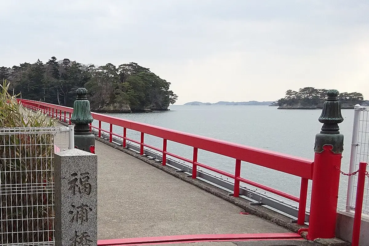The vermilion railing of Fukuura Bridge running out across the water to the pine-covered island of Fukuurajima, with a stone pillar at the entrance