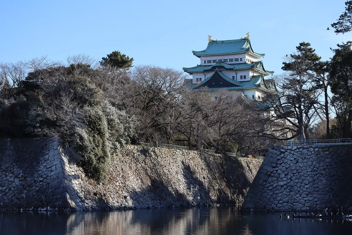 Across the water of the moat: the closed main keep of Nagoya Castle standing above its stone walls, with two golden shachihoko on the roof ridge