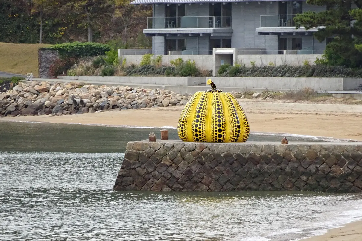 Yayoi Kusama's yellow polka dot pumpkin standing alone at the end of a stone pier by the sea on Naoshima, with the beach and a low concrete building behind it