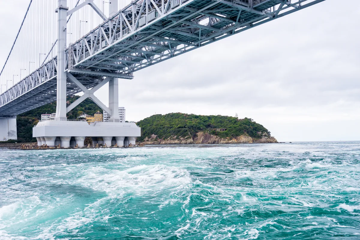 The Onaruto Bridge seen from the water, with churning turquoise current directly beneath the pylon and a green island behind