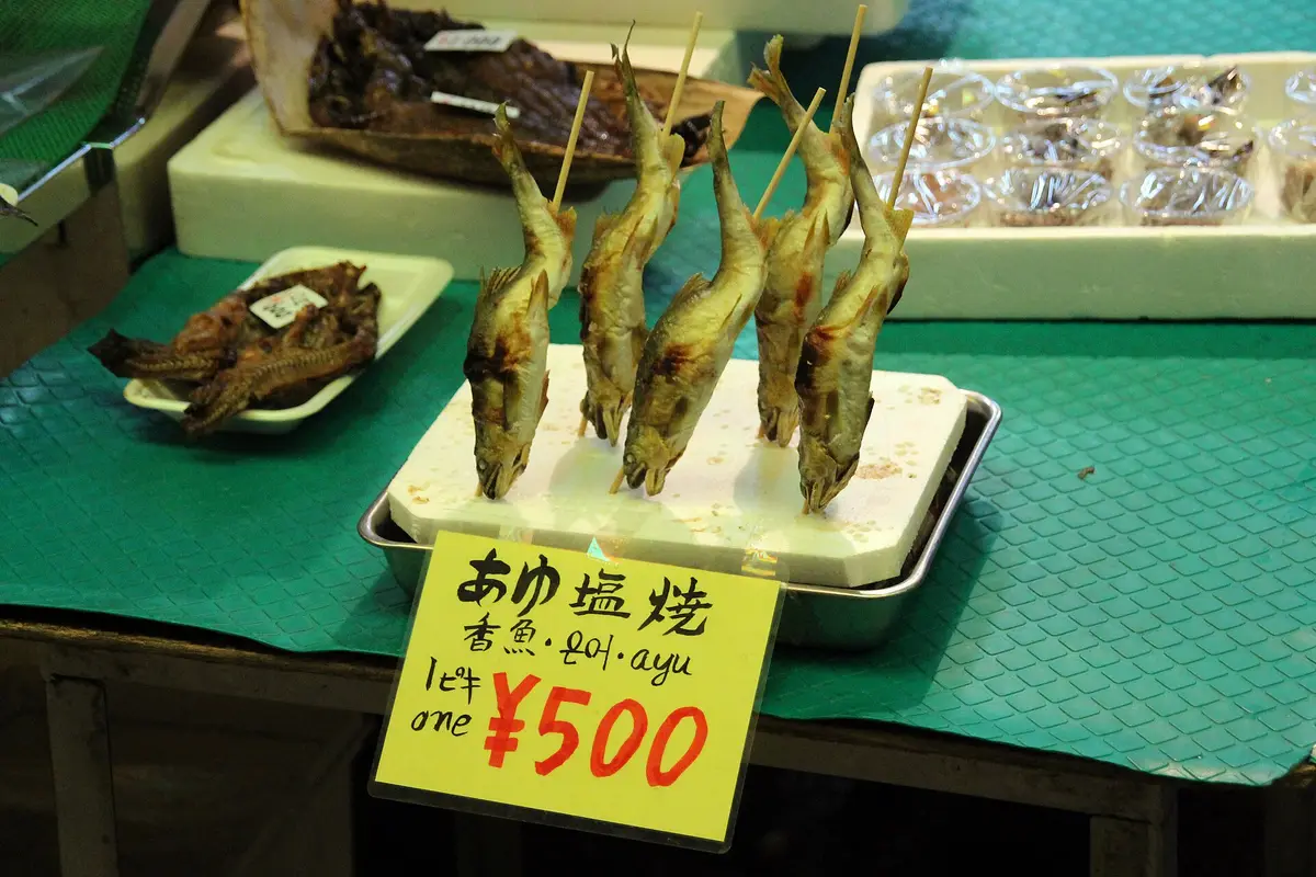 Skewers of salt-grilled ayu fish standing on a tray at a Nishiki Market stall counter, with a handwritten price card in front