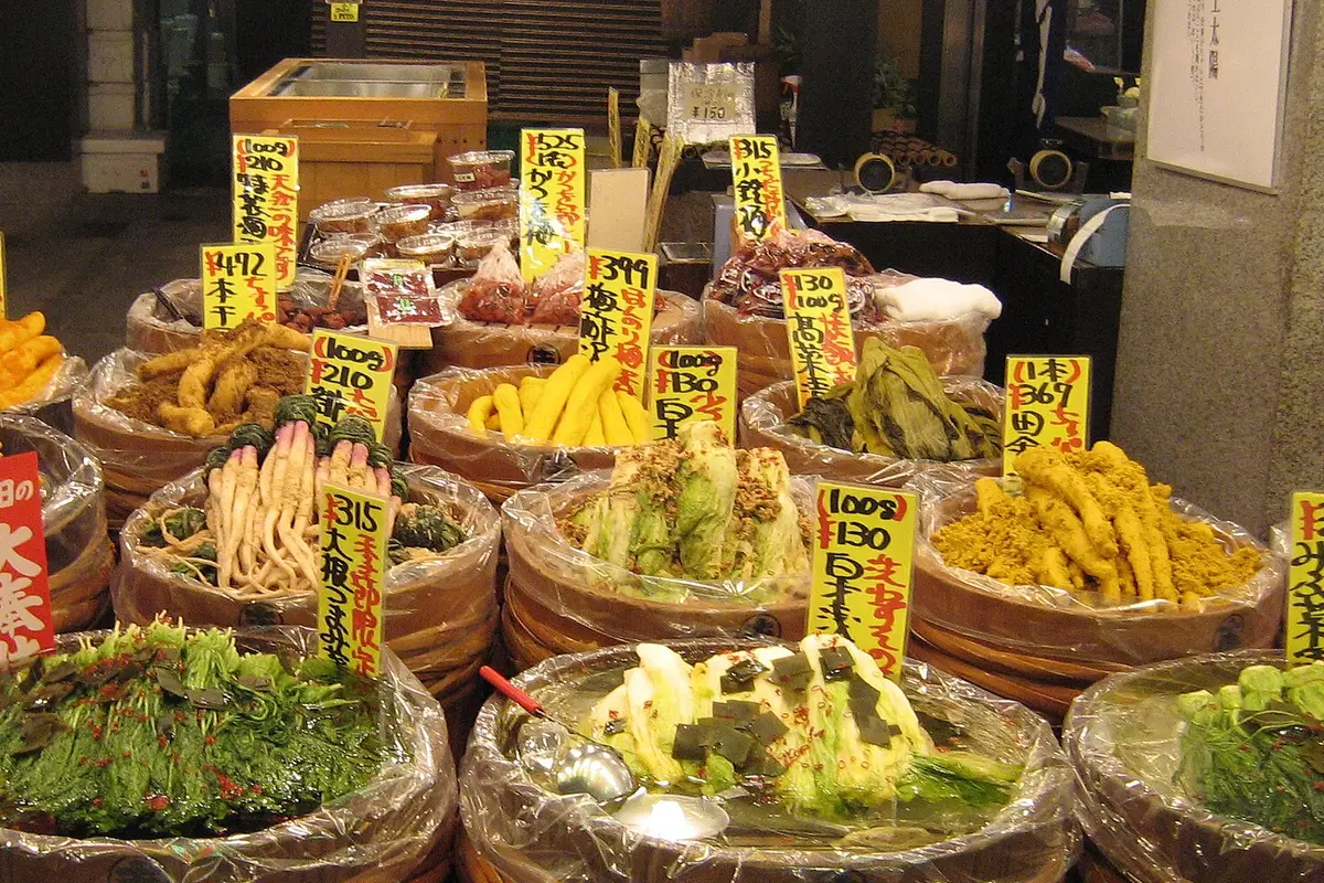 Rows of wooden tubs of Kyoto pickles at a Nishiki Market shopfront, each with a handwritten card giving the price by weight