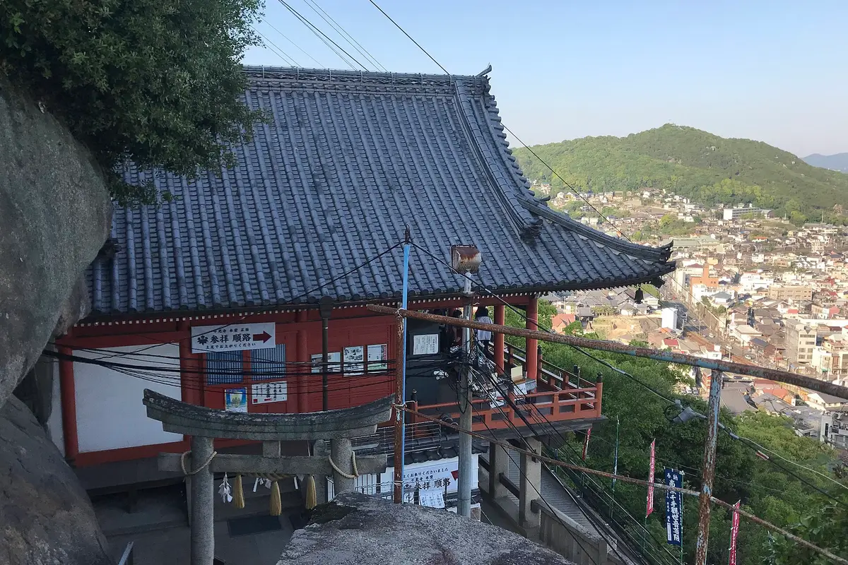 Seen from the path above it: the tiled roof and vermilion veranda of Senkoji's main hall, built out on pillars against the rock, with the town of Onomichi below