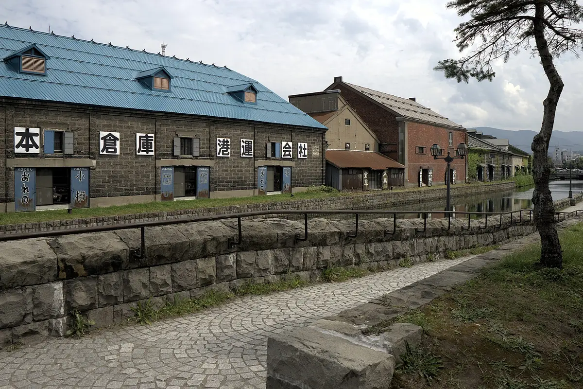 Seen from the walkway: a row of old stone warehouses along the Otaru Canal on a summer afternoon, a gas lamp and the water on the right