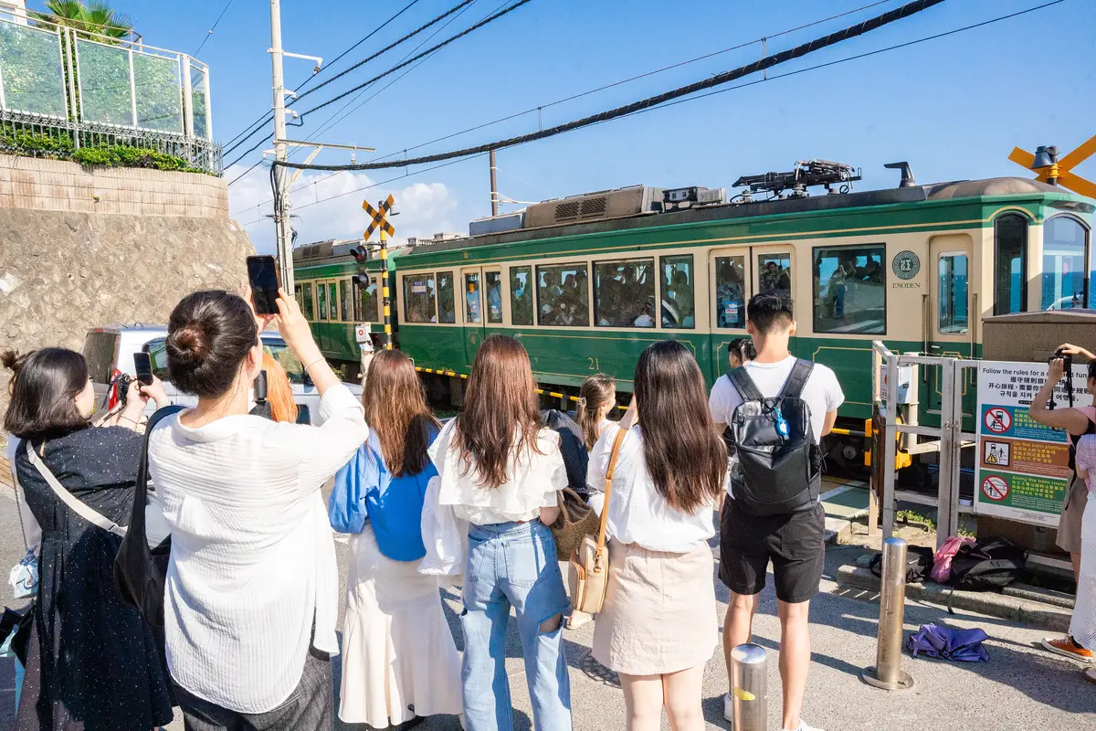 A row of visitors seen from behind, phones raised, watching a green train pass the seaside level crossing at Kamakura-Kōkō-Mae, beside a multilingual sign