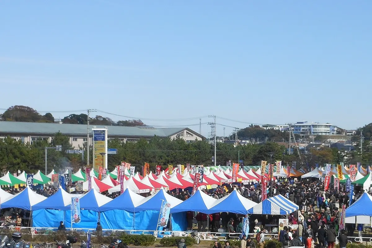 Rows of festival stalls with tall banners on an open ground in Oarai, with a dense crowd filling the lanes between them