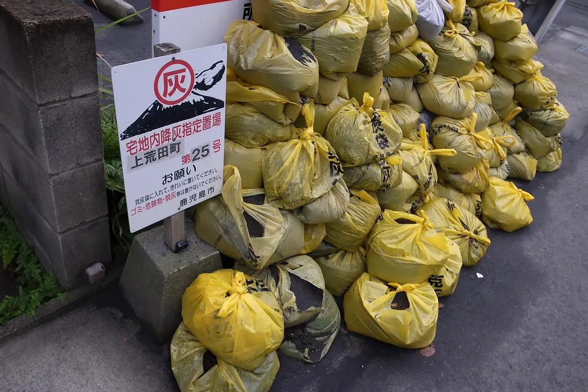 Yellow ashfall bags stacked at a designated collection point on a Kagoshima street, next to a city sign showing an erupting volcano
