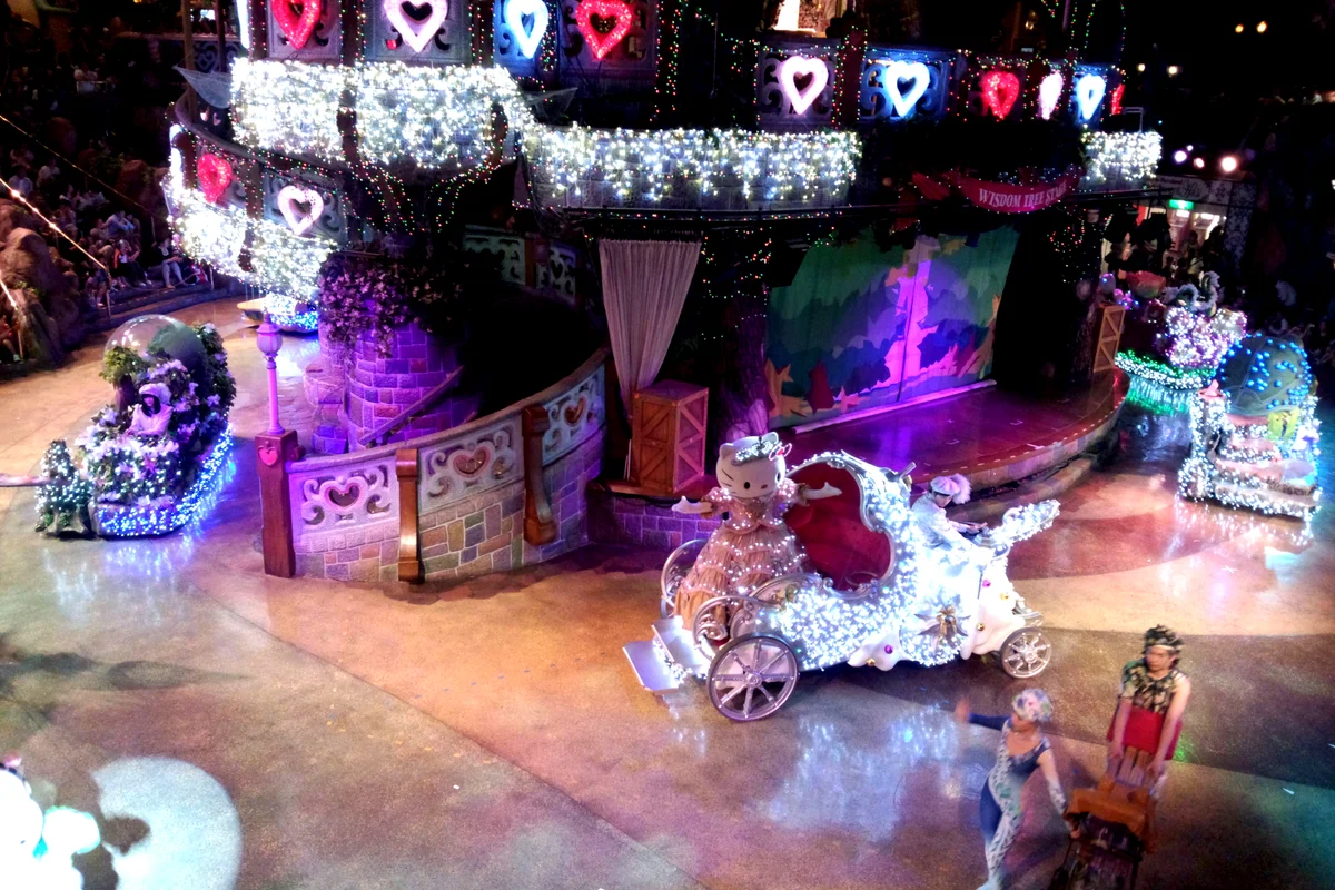 Hello Kitty riding a lit-up carriage float during an evening parade at Sanrio Puroland, with an illuminated stage behind her